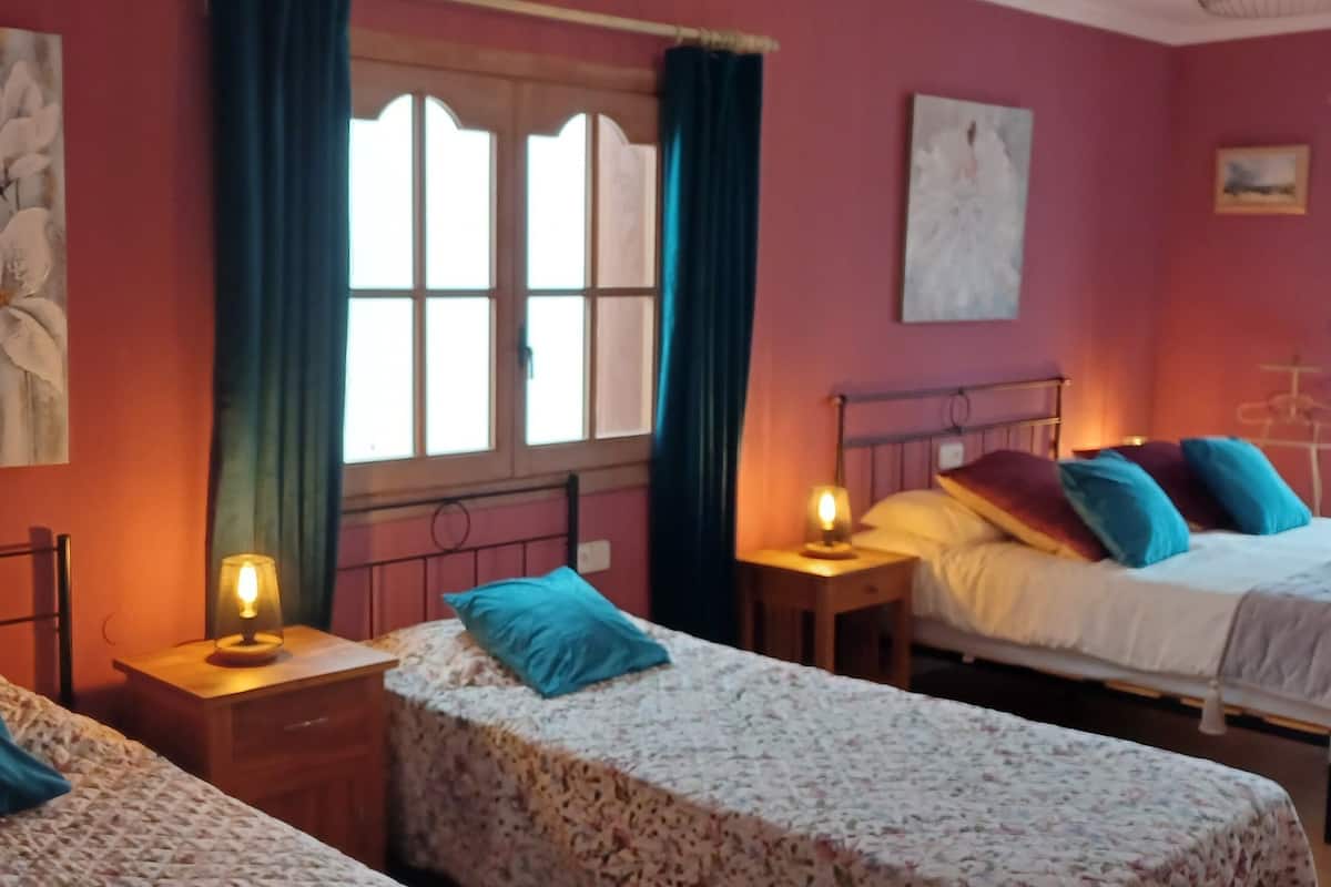 A room with three beds, each with a different pillow color, a window with curtains, and wall art.