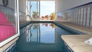 Una piscina interior con agua cristalina, paredes de azulejos y vistas a plantas y flores al aire libre.