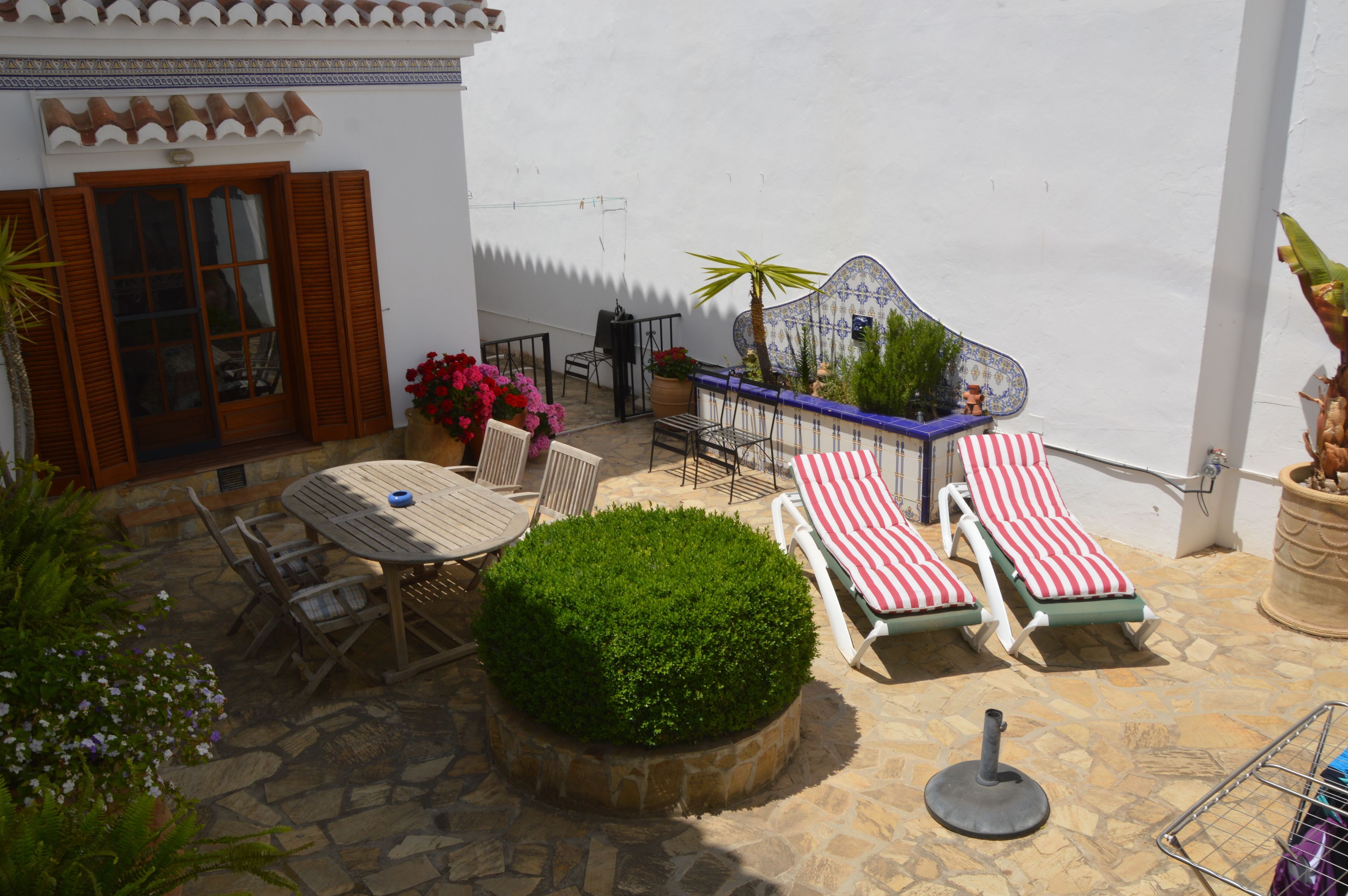 A patio with a round table and chairs, two striped lounge chairs, a small potted plant, and a decorative wall piece.
