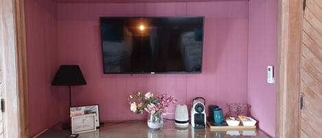 A room with a flat-screen TV mounted on the wall, a lamp, a vase with flowers, and a table with a kettle, cups, and a tray of bread.
