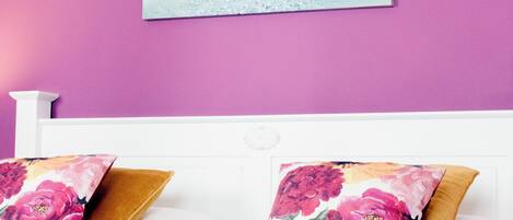 Quarto com cabeceira branca, almofadas florais e cobre-leito.