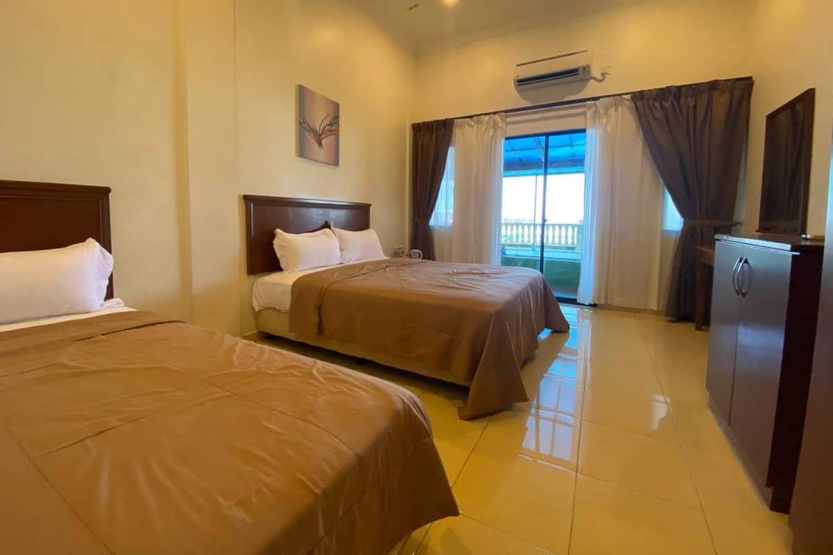 A hotel room with two beds, a ceiling fan, an air conditioning unit, a small refrigerator, and a window with curtains.