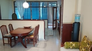 A dining area with a table and chairs, a sofa with patterned cushions, a water dispenser, and a ceiling fan. - Qua Bruce Apartment (Diani Beach)