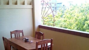 A dining area with a wooden table and chairs, a window with a view of greenery and buildings, and a white shelf unit on the wall. - Qua Bruce Apartment (Diani Beach)