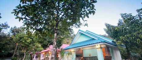 A small, single-story building with a blue roof and light green walls, surrounded by lush greenery and trees.