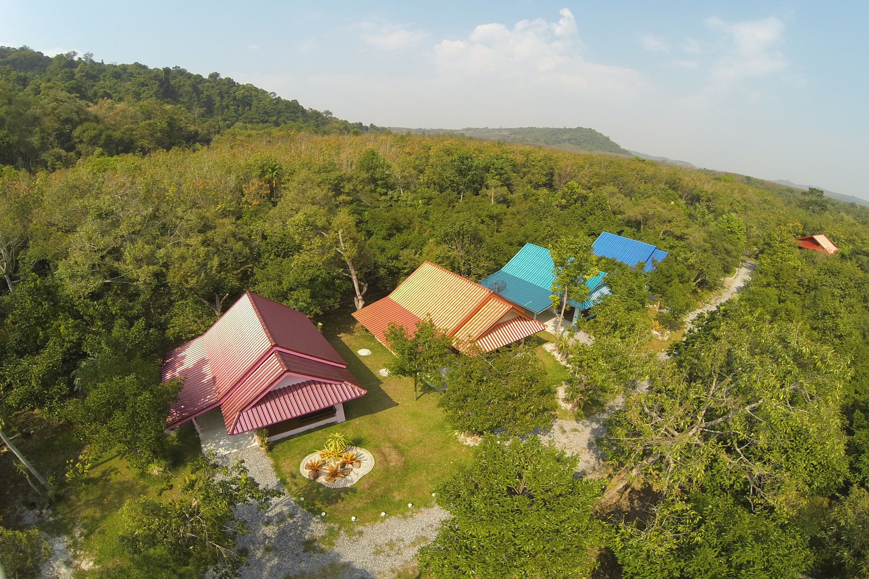 A cluster of buildings with red and blue roofs surrounded by dense greenery and hills in the background.