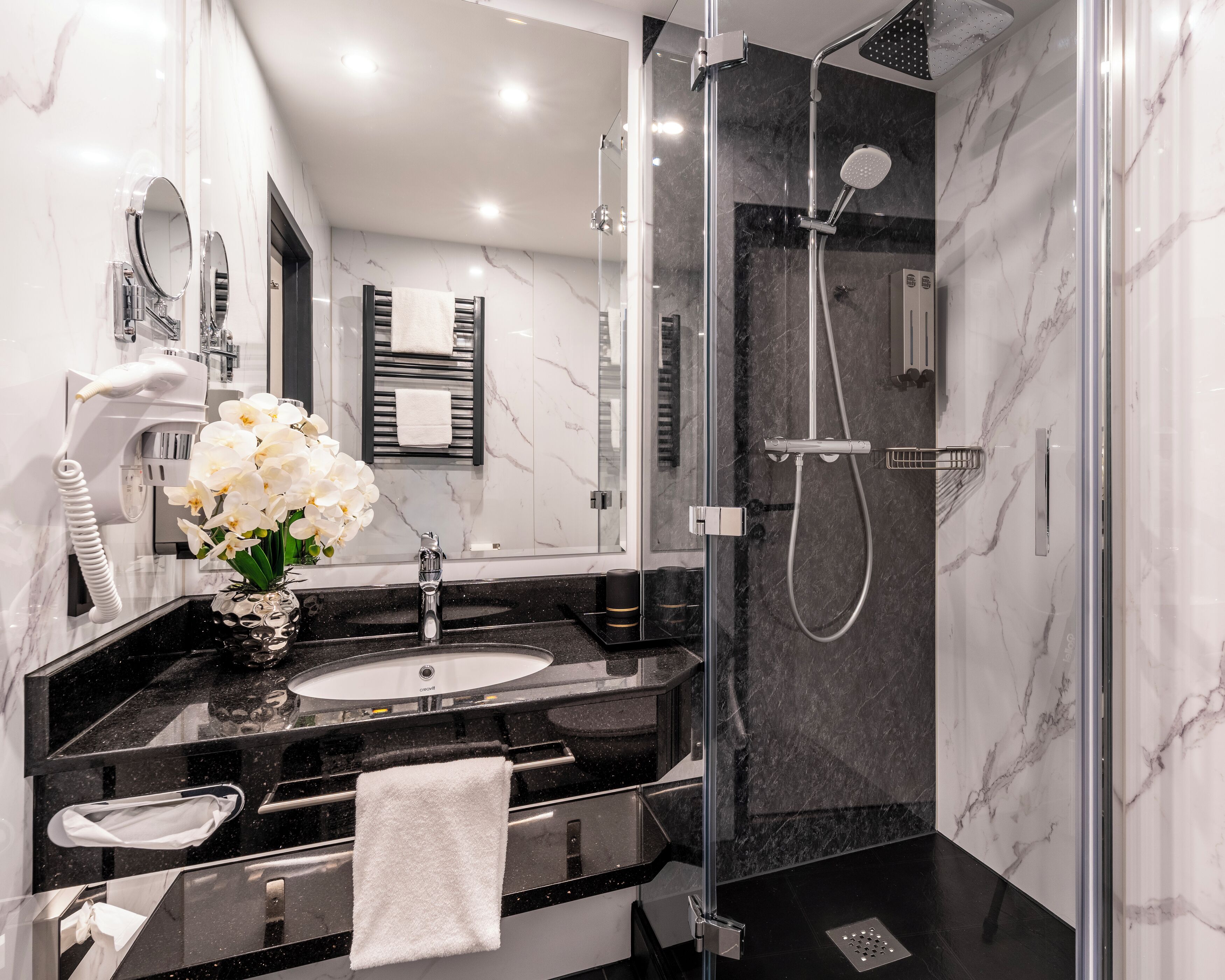 A modern bathroom with a glass-enclosed shower, a sink with a marble countertop, and a vase with white flowers.