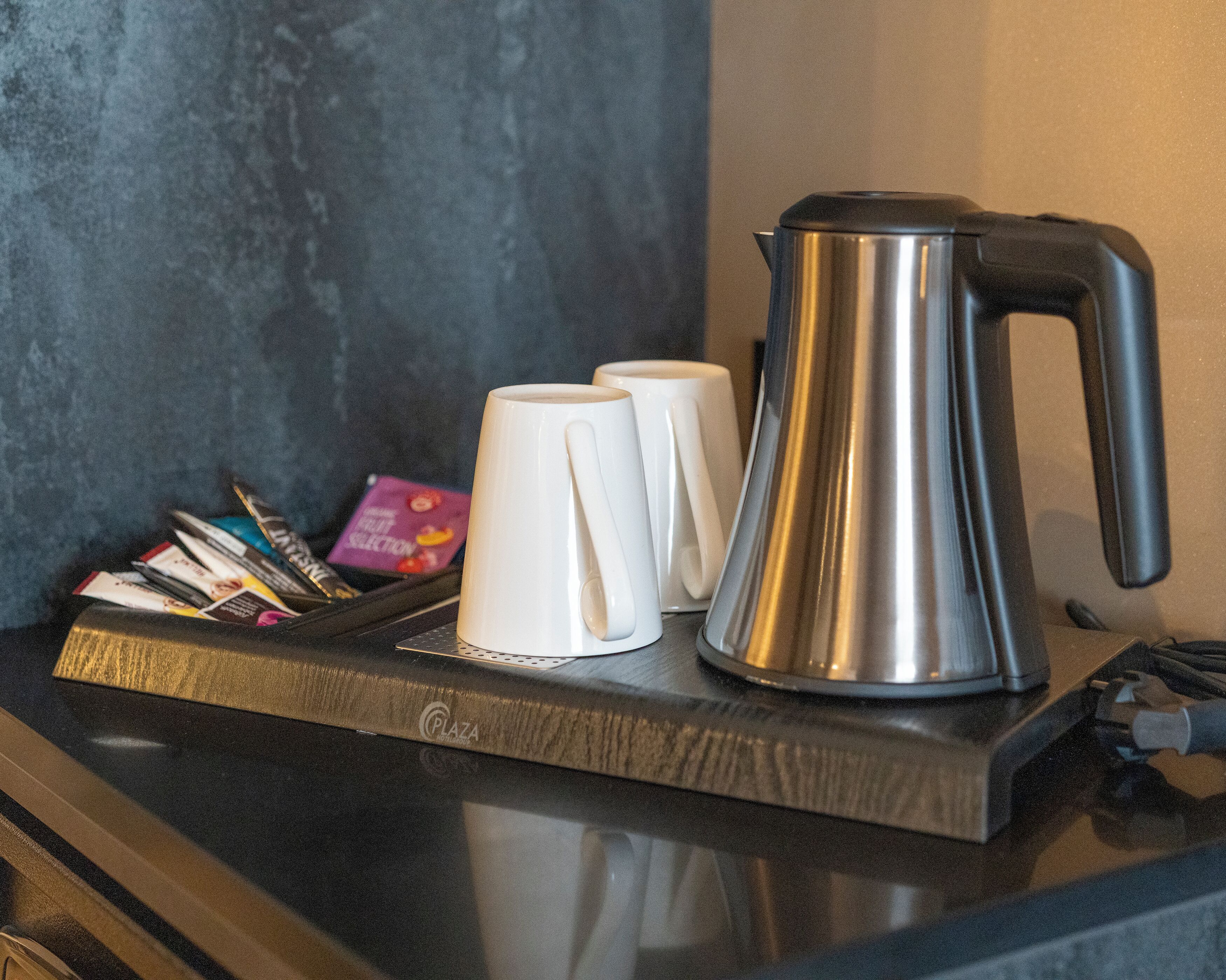 Uma bancada de cozinha com uma cafeteira, duas canecas brancas e uma bandeja com revistas e uma caixa cor-de-rosa.