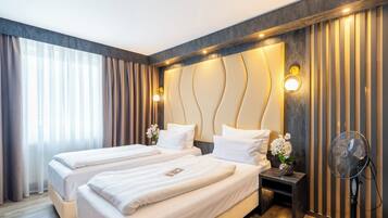 Habitación de hotel con dos camas, una mesita de noche, un ventilador de pared y un ventanal con cortinas.