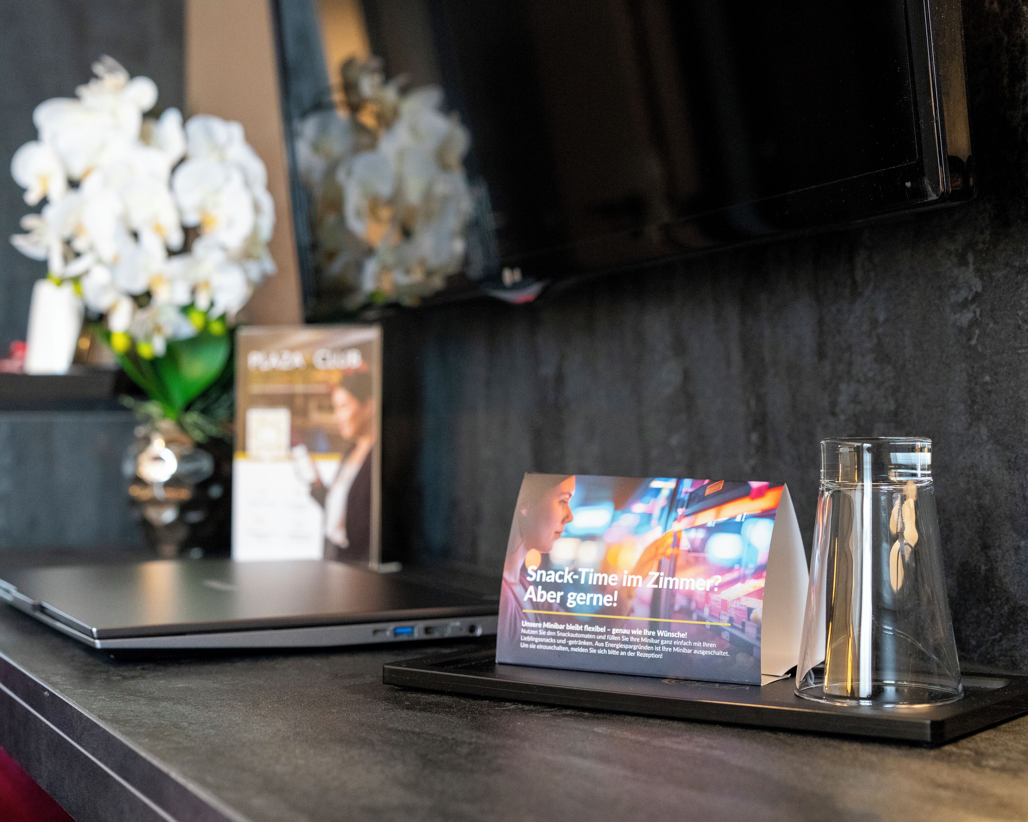 Un ordinateur portable posé sur un bureau, à côté d’un verre d’eau, d’une affiche avec du texte et d’un vase rempli de fleurs blanches.