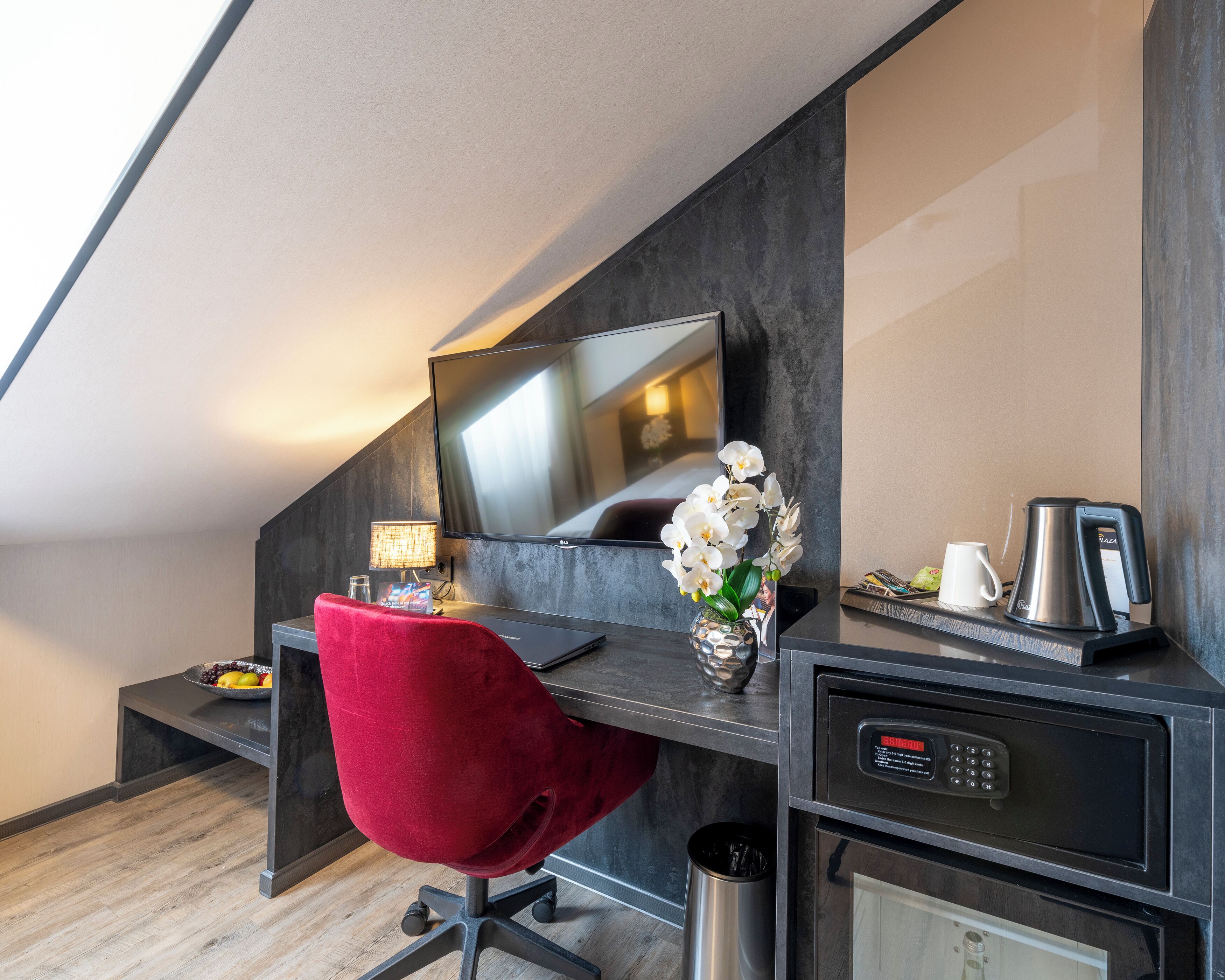 A modern hotel room with a desk, a red chair, a flat-screen TV, a coffee maker, and a trash can.