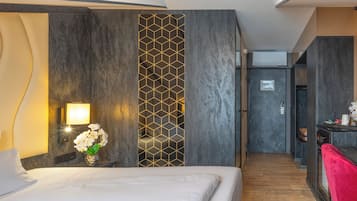 A hotel room with a bed, a nightstand with a lamp, a decorative wall panel, and a view of the hallway.