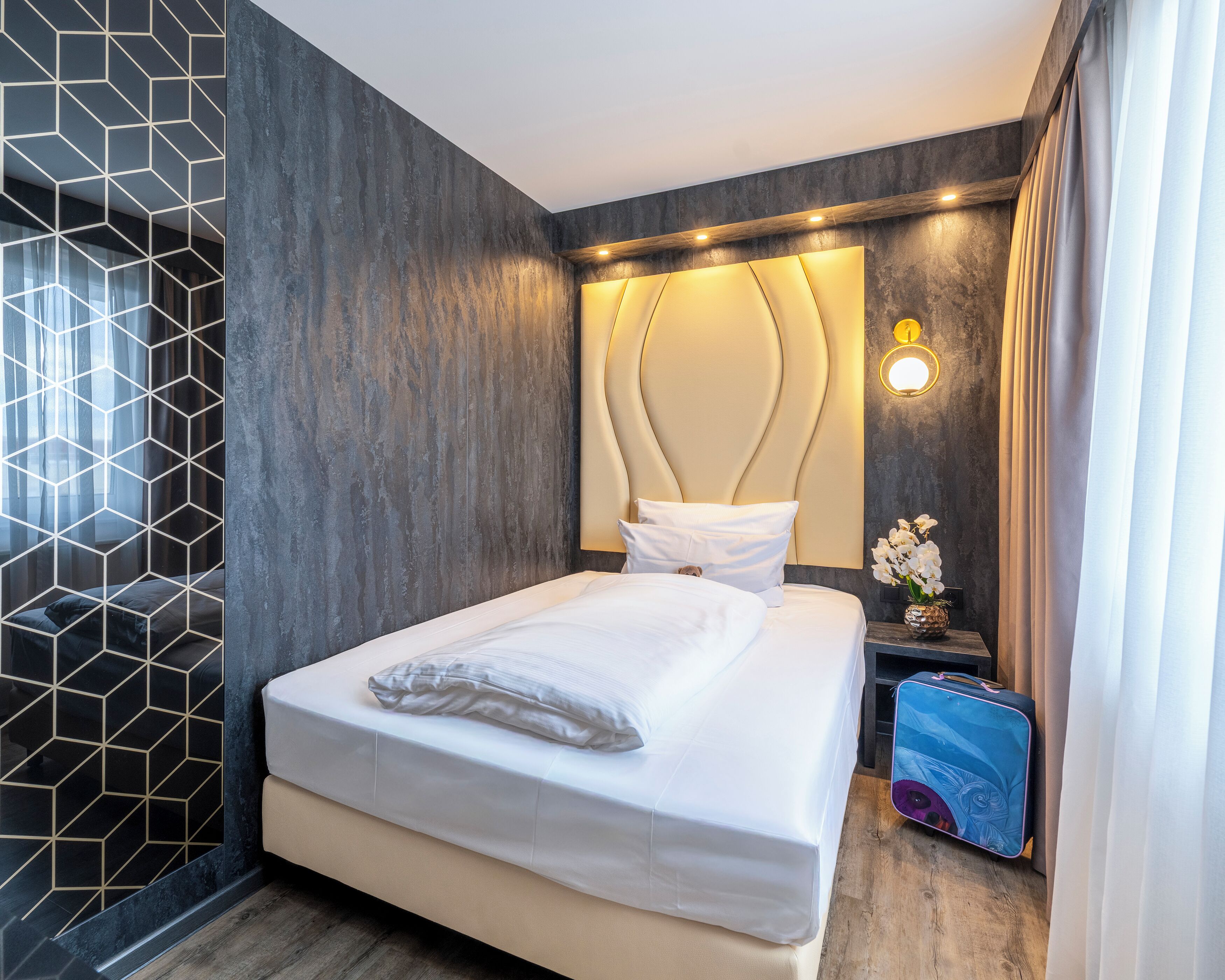 A modern hotel room with a geometric-patterned wall, a large bed with white bedding, a headboard with integrated lighting, a nightstand with a vase of flowers, and a suitcase on the floor.