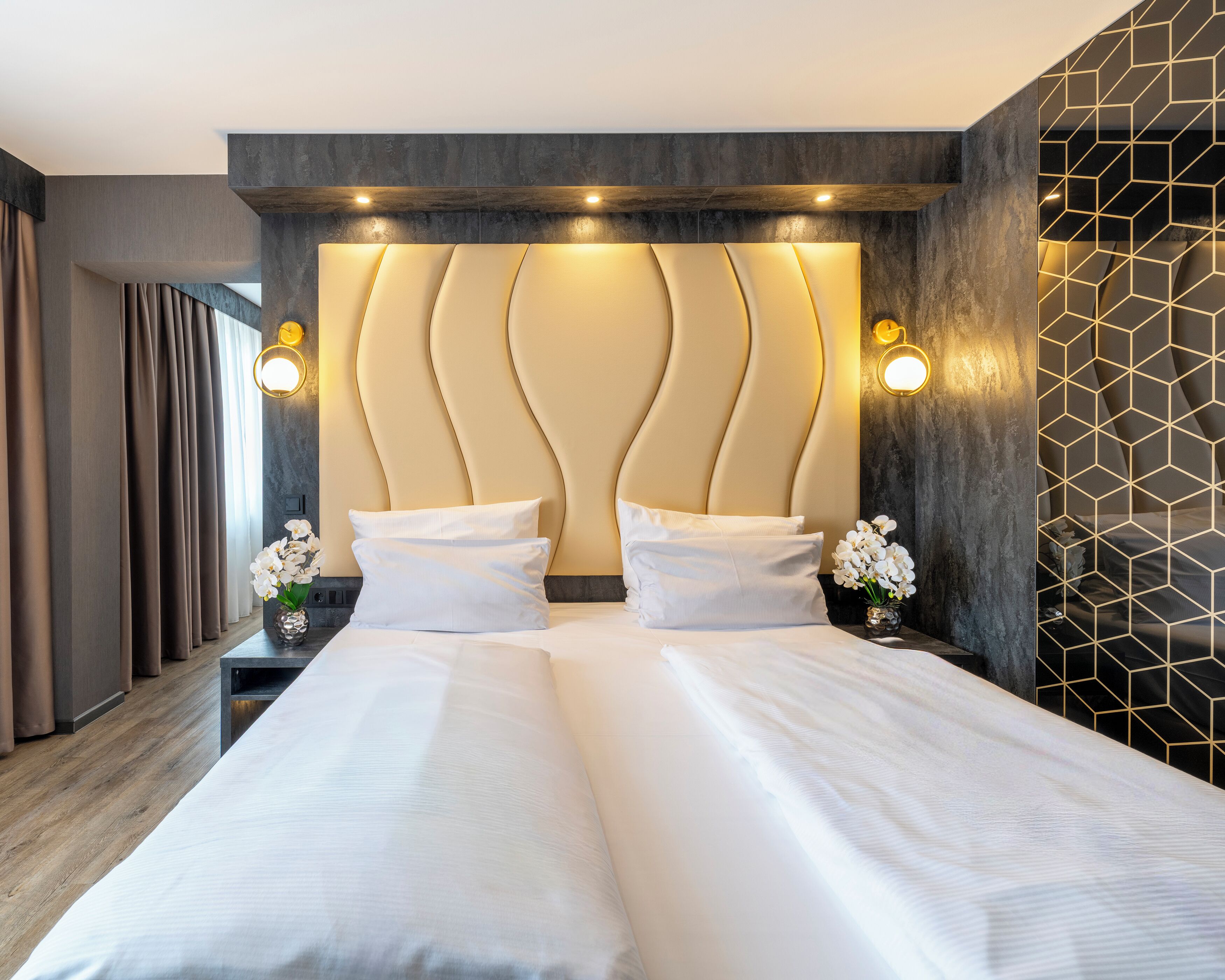 Een tweepersoonsbed met een modern hoofdeinde, nachtkastjes met lampen, en een geometrisch patroon op de muur.