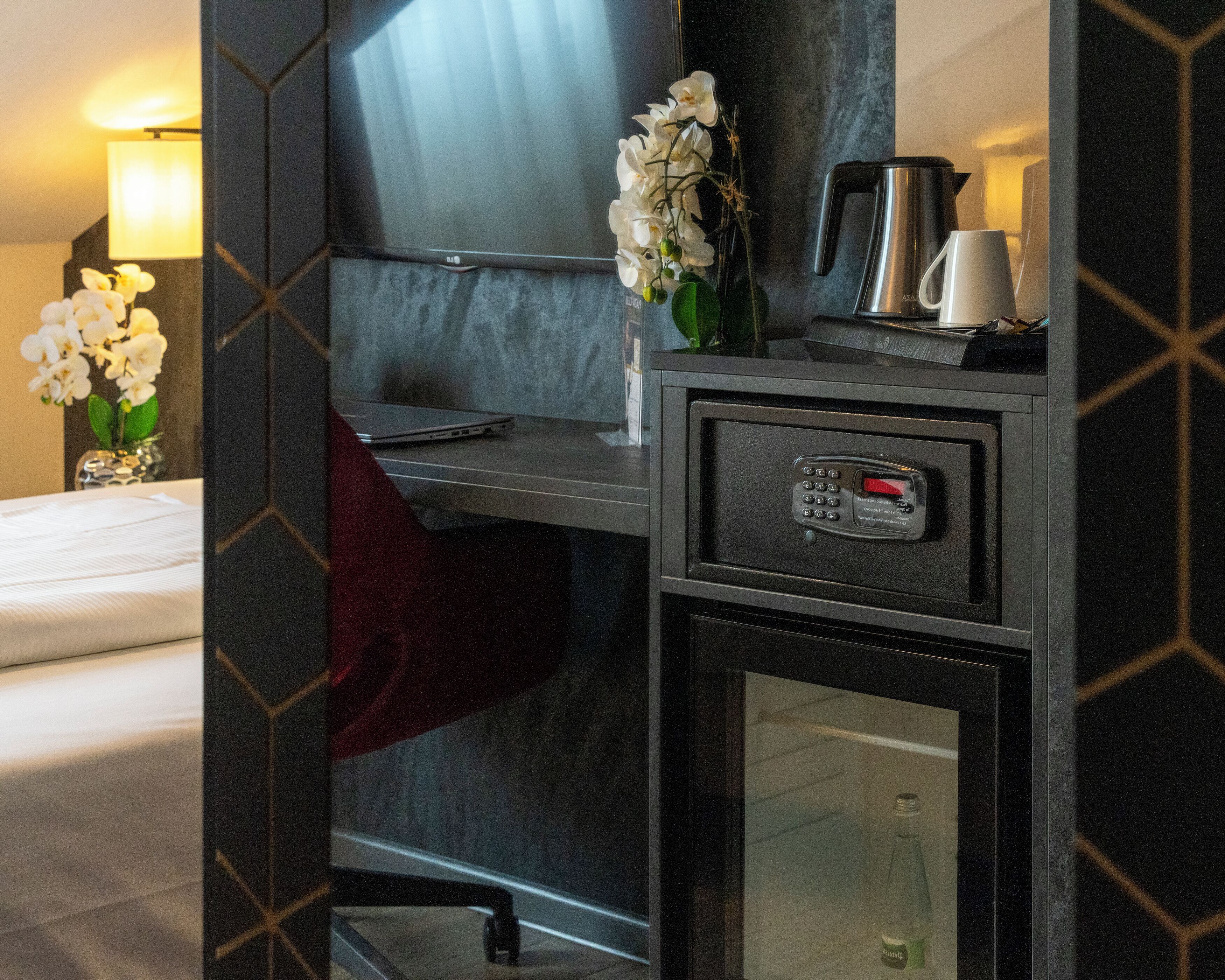 A hotel room with a bed, a desk with a laptop, a coffee maker, a minibar, and a wall-mounted TV.