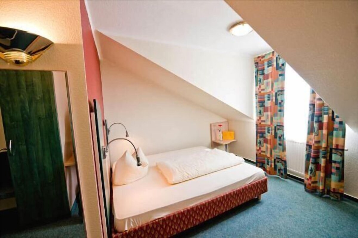 A small, single-bed room with a sloped ceiling, a bedside table with a lamp, and colorful curtains.