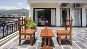 A balcony with two wooden tables and chairs, a view of a mountain, and a building with large windows and air conditioning units. - The White Lake Hotel (Tansen)