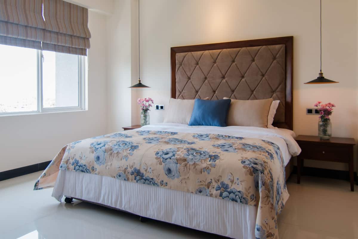 Une chambre à coucher bien rangée, avec un grand lit, une tête de lit, des tables de chevet et une fenêtre avec des rideaux.