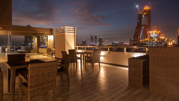 A rooftop terrace with wicker furniture, a dining table, and a view of the city skyline at dusk. - Rococo Residence (Colombo)