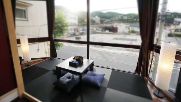 Ein Zimmer mit Tatami-Matten, einem niedrigen Tisch und Kissen. Ein großes Fenster bietet Blick auf einen Parkplatz und Gebäude.