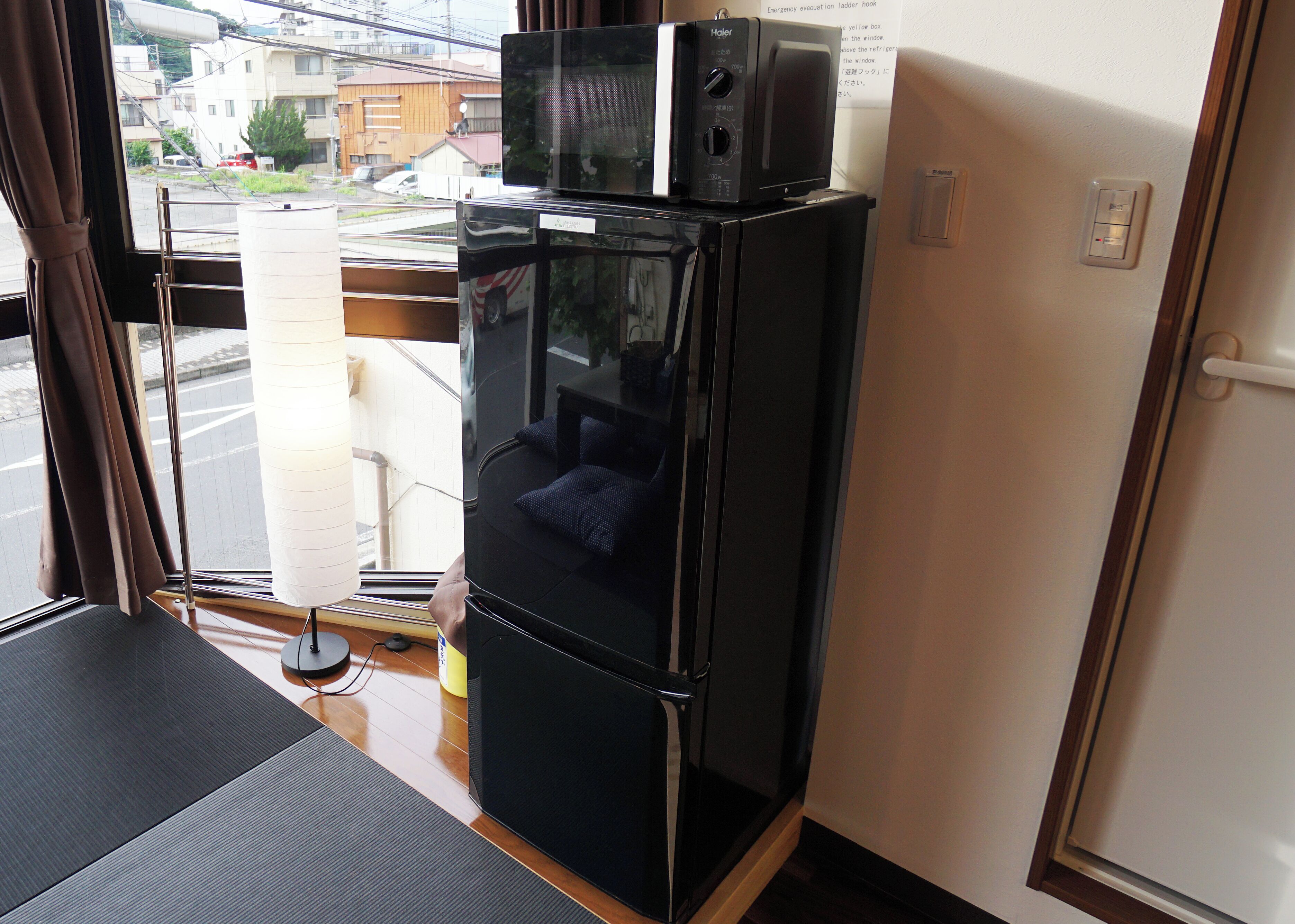 A compact kitchenette with a microwave and refrigerator, a window with a view of buildings, and a door with a light switch.
