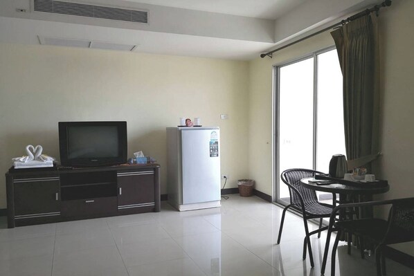 A hotel room with a flat-screen TV, a minibar, a dining area with a table and chairs, and a large window with curtains.