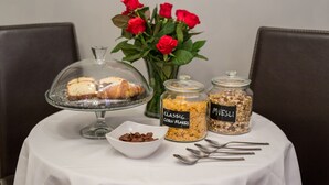 Un tavolo con una torta su un piedistallo in vetro, due barattoli etichettati “Classic Corn Flakes” e “Muesli”, una piccola ciotola di gocce di cioccolato e un vaso di rose rosse.