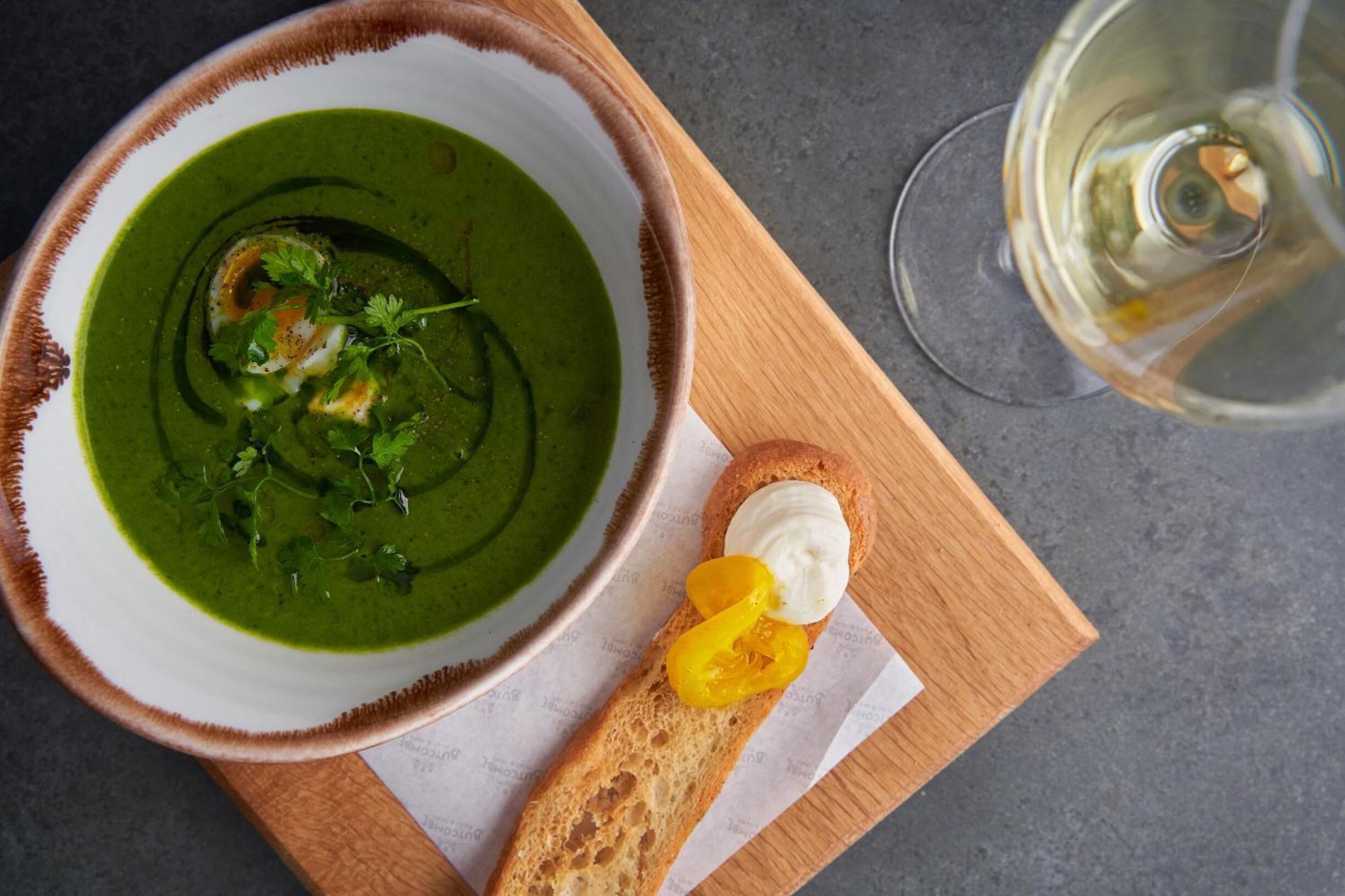 A bowl of green soup with a garnish, a slice of bread with a dollop of cream and a slice of yellow fruit, and a glass of white wine on a wooden board.