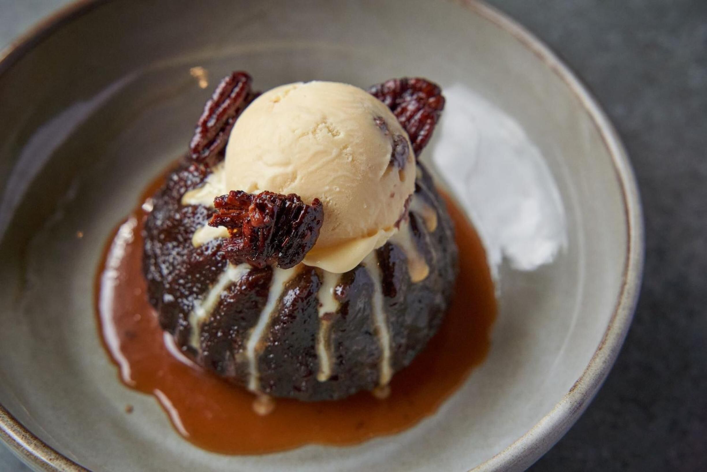 A dessert consisting of a chocolate lava cake with caramel sauce and a scoop of vanilla ice cream, garnished with pecans.