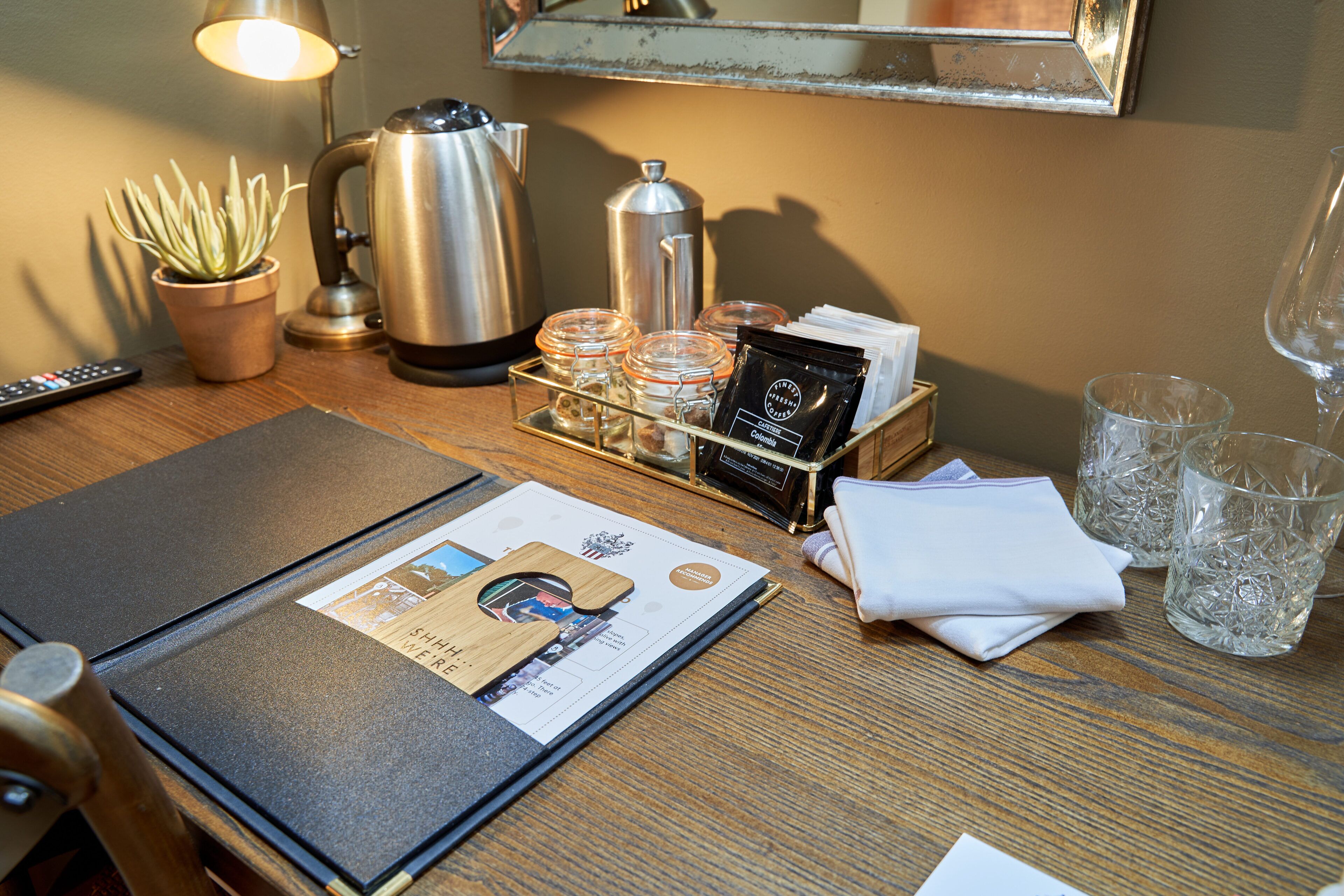 Habitación de hotel ordenada con una mesa de madera, cafetera, hervidor, planta, bandeja con frascos de vidrio, menú y control remoto.