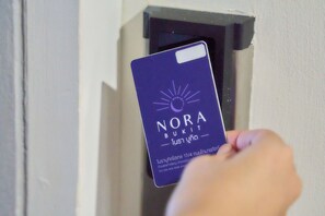 Eine Hand hält eine Karte mit dem Text 'NORA BUKIT' und einem Sonnensymbol, die in eine Aussparung in einer Wand eingeführt wird.