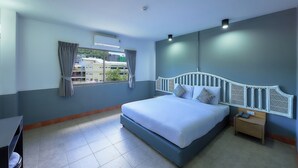A bedroom with a large bed, a nightstand, a window with curtains, and a view of buildings outside. - Nora Bukit Hotel (Phuket)