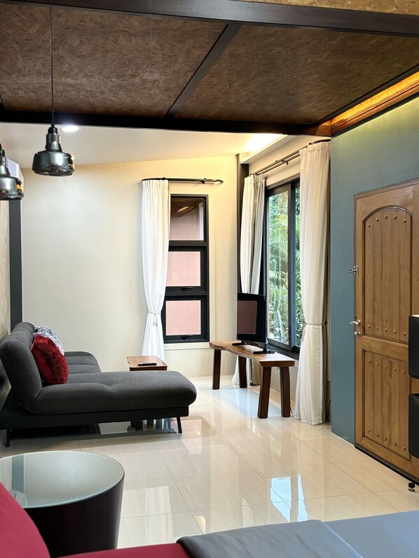 A modern living room with a sofa, a TV on a wooden stand, and a large window with curtains. - Cafe'@luv22 (Wichit)
