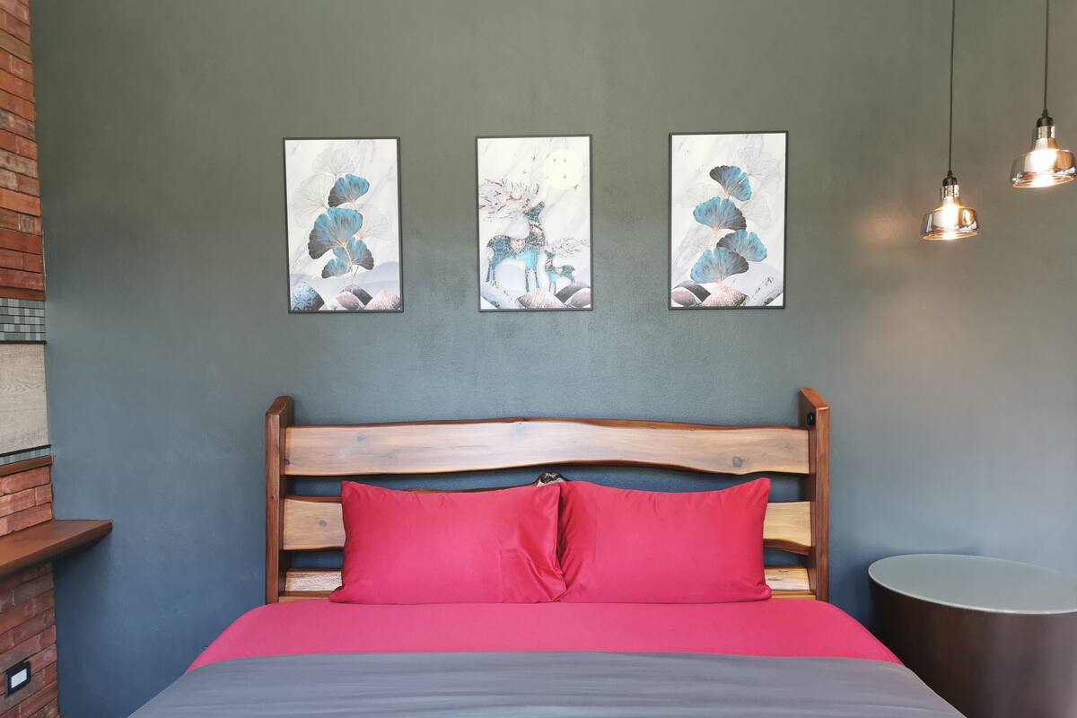 Un dormitorio con cabecera de madera, almohada roja y colcha gris. Hay tres cuadros enmarcados en la pared y dos lámparas colgantes.