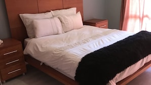 Une chambre à coucher avec une tête de lit en bois, un lit recouvert de linge de lit blanc, un plaid noir et une table de chevet en bois dotée de tiroirs.