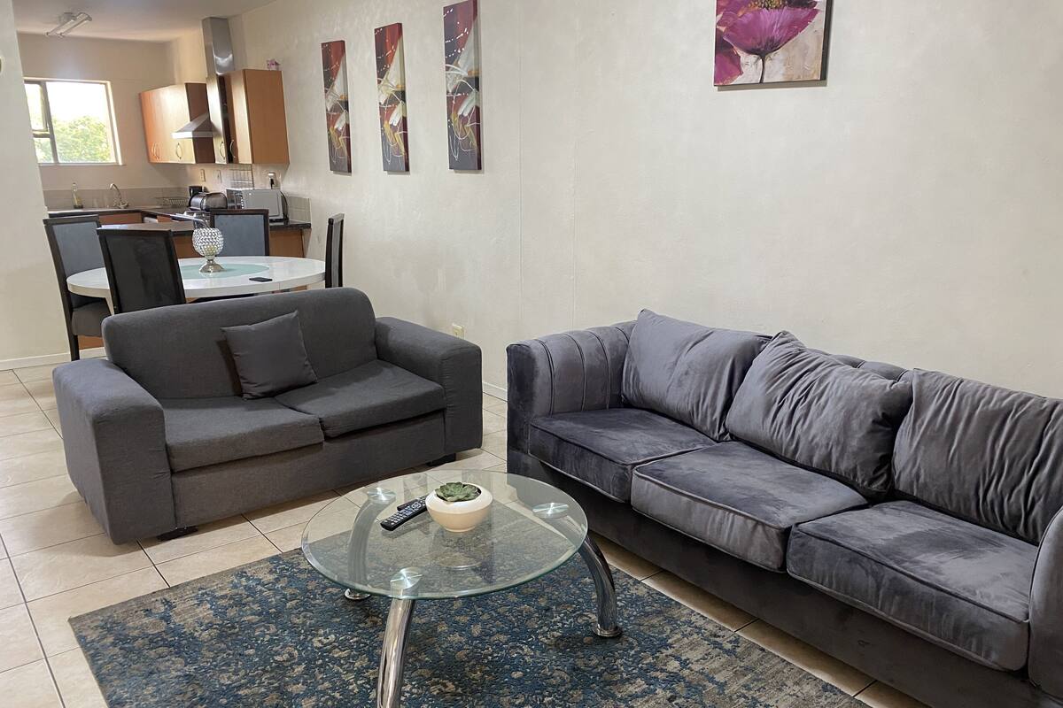 A modern living room with two grey sofas, a glass coffee table, and a blue patterned rug. A kitchen with wooden cabinets and a dining area with a table and chairs is visible in the background.