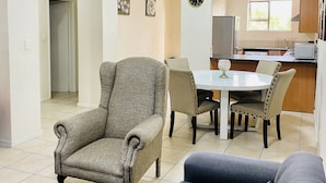 Un salon avec un canapé, un fauteuil et une table à manger. Au fond, on aperçoit une cuisine équipée d’un réfrigérateur, d’un four  à micro-ondes et d’un évier.