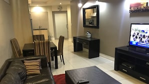 A living room with a black leather sofa, a red rug, a coffee table, a TV, a dining table with chairs, a black desk, and a mirror on the wall. - First Divine Suites - Hydro Park (Sandton)