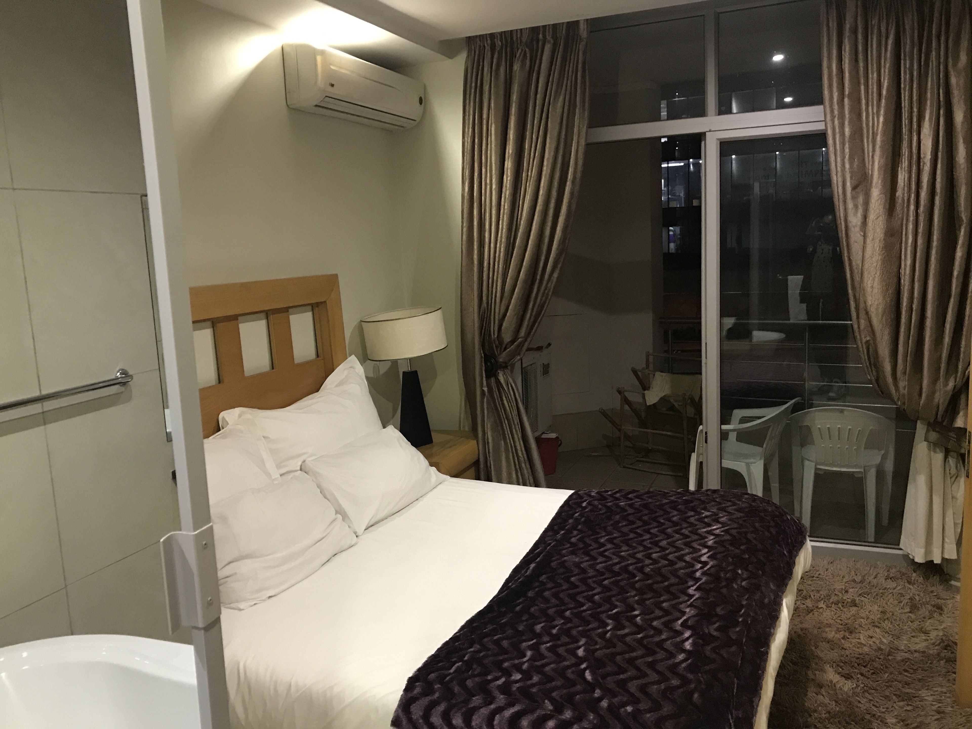 Une chambre d’hôtel comprenant un lit, une table de chevet, une lampe, un appareil de climatisation, une fenêtre avec des rideaux et une vue sur le balcon extérieur.