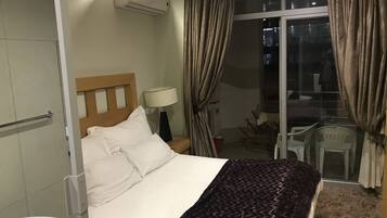 Une chambre d’hôtel comprenant un lit, une table de chevet, une lampe, un appareil de climatisation, une fenêtre avec des rideaux et une vue sur le balcon extérieur.