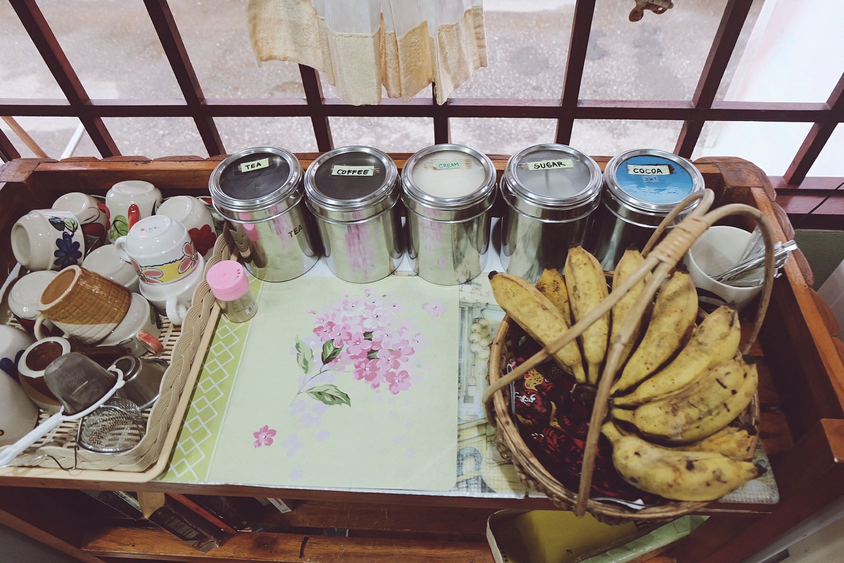 A tray with tea, coffee, cream, sugar, and cocoa containers, a floral tablecloth, a strainer, a basket of bananas, and various cups and mugs.