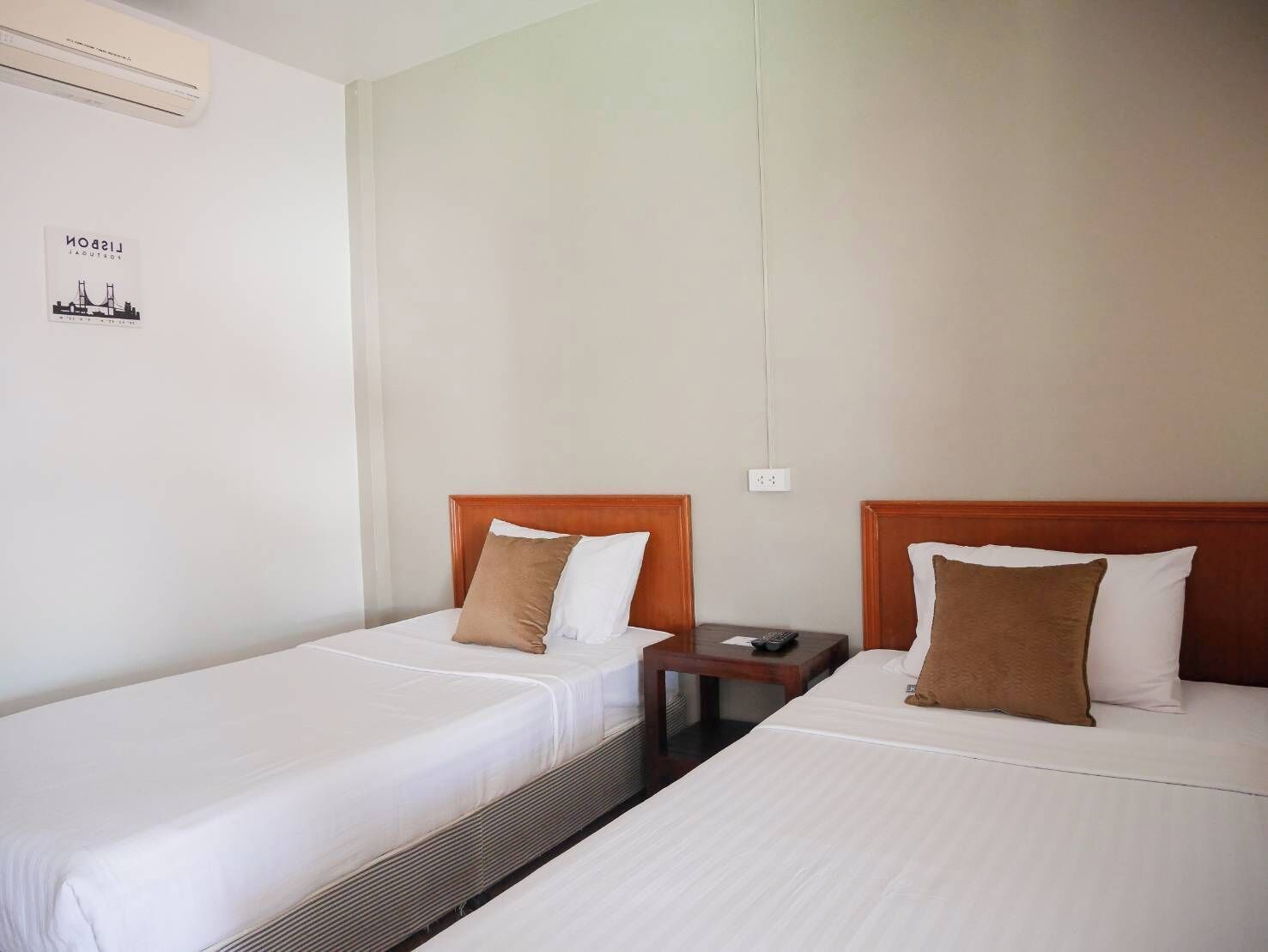 A hotel room with two single beds, each with a wooden headboard, white bedding, and a brown pillow. There is a small wooden nightstand between the beds with a remote control on it.