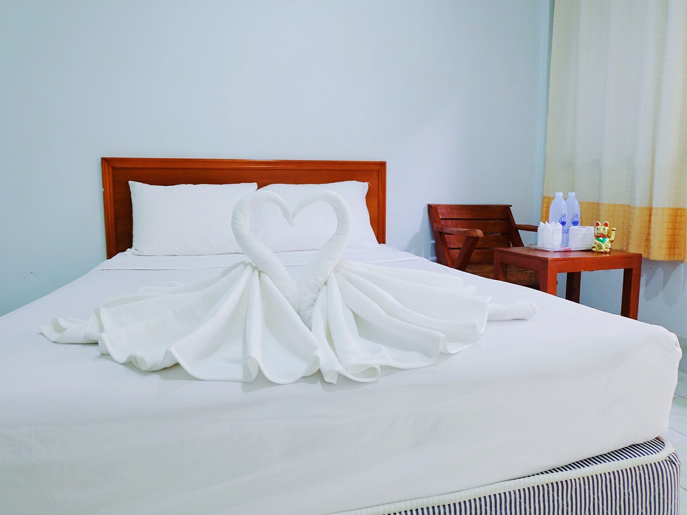 Habitación de hotel con cama bien tendida, cabecera de madera, una silla de madera, una mesita con artículos de aseo y una ventana con cortinas.