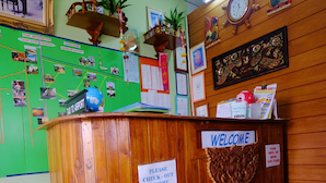 A wooden reception desk with a 'Welcome' sign, a 'Taxi to Airport' sign, and various informational posters and notices. - Orchid House (Chiang Rai)