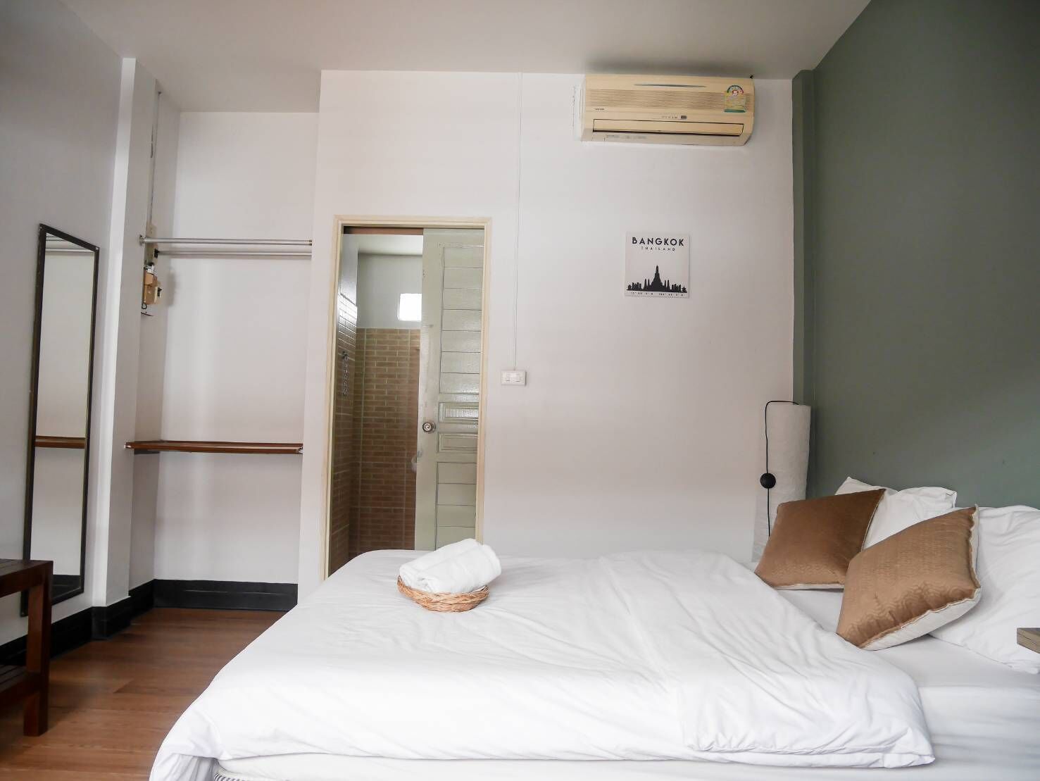 Hotellihuone, jossa on sänky, puinen tuoli, pyyhe sängyllä, seinään asennettu ilmastointilaite ja ovi, joka johtaa kylpyhuoneeseen.