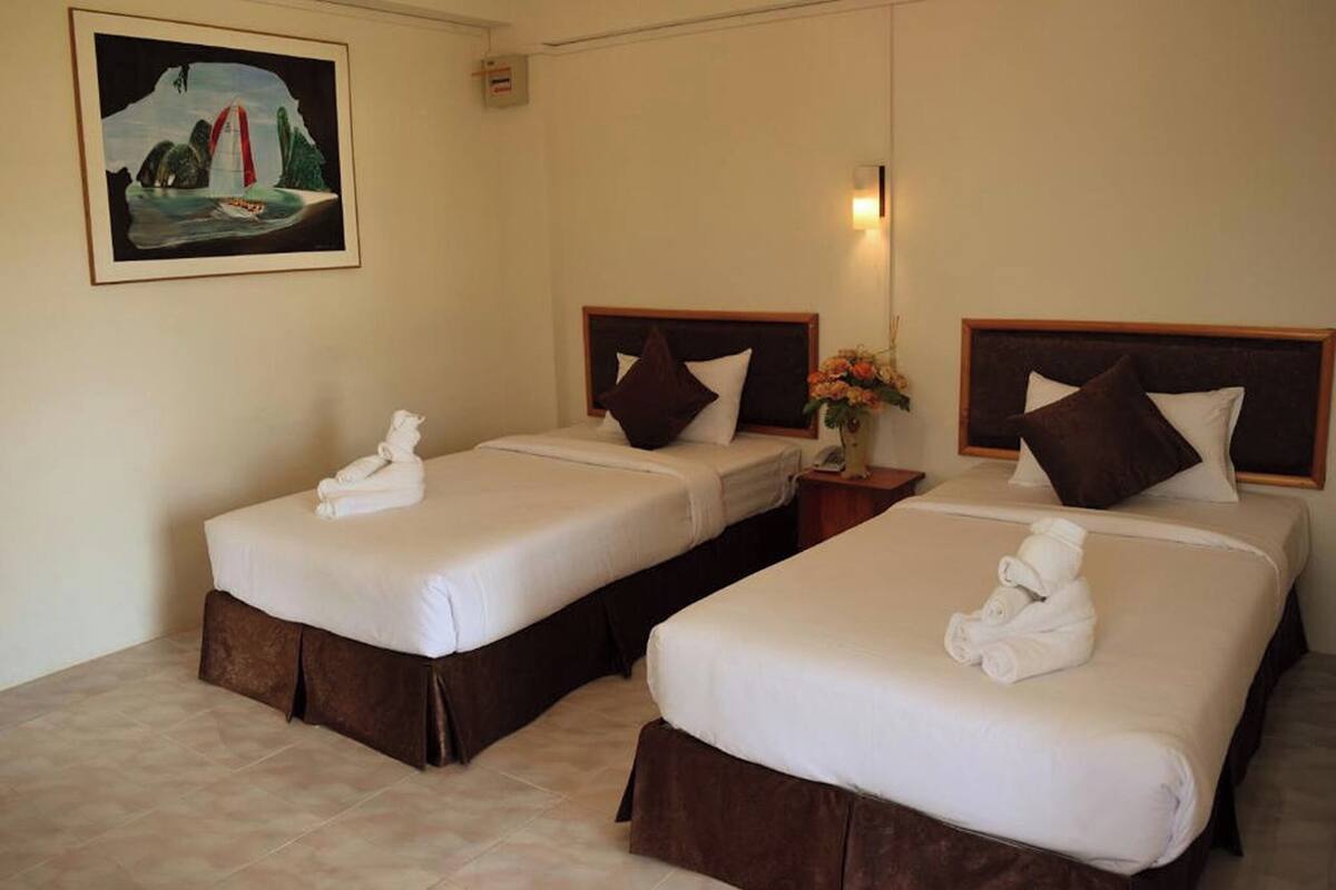 A hotel room with two beds, each with a brown headboard and white bedding, a small bedside table with a vase of flowers, and a framed picture on the wall.