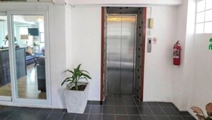 An elevator in a hallway with a potted plant, a fire extinguisher, and a glass door leading to a room. - Hotel De Ratt (Ratsada)