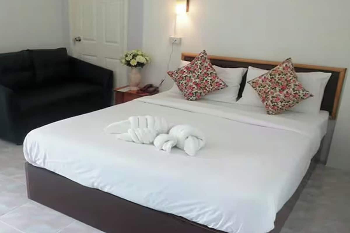 A neatly made bed with white linens and decorative pillows, a dark-colored sofa, and a small table with a vase of flowers.