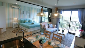 A modern bedroom with a large bed, a sitting area with a chair, a dining table with chairs, and a balcony with a view. - Rain Cha-am Hua-hin by Noppawan (Cha-am)