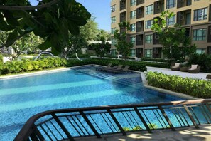 A swimming pool with lounge chairs, surrounded by greenery and a multi-story building.